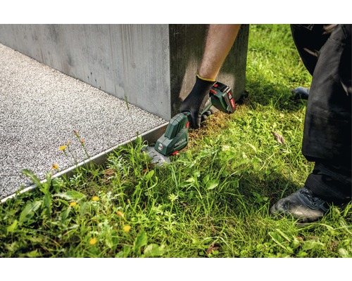 Zahradník stříhá trávu akumulátorovými nůžkami na trávu značky Metabo podél betonové zdi.