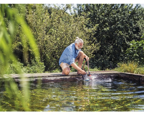 Muž klečí na okraji jezírka a čistí jezírko pomocí vysavače kalu.