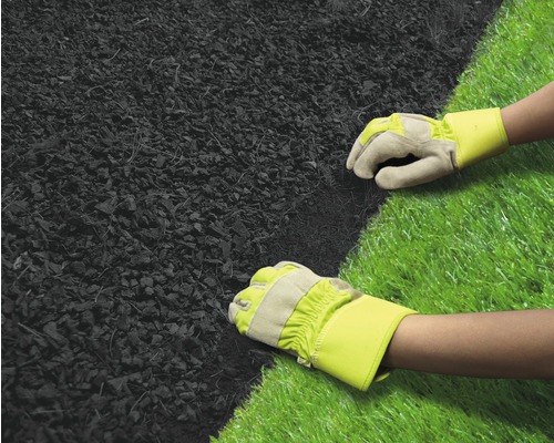 Dvě ruce v pracovních rukavicích pokládají mulč vedle umělého trávníku.