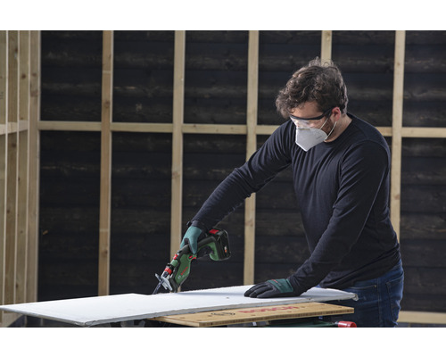 Muž řeže sádrokartonovou desku akumulátorovou přímočarou pilou a má ochranné brýle a obličejovou masku.