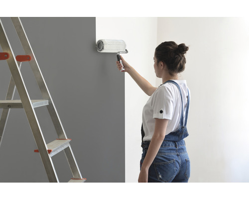 Žena maluje zeď válečkem na barvu, vedle stojí žebřík