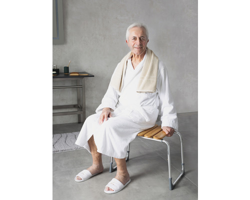 Muž v županu sedí na sprchové stoličce s dřevěnými lamelami a kovovým rámem.