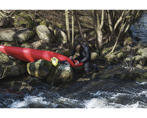 Muž opravuje kajak na břehu řeky s kameny a stromy.