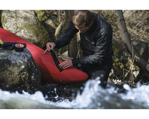 Muž opravuje červený kajak černou lepicí páskou poblíž skal a tekoucí vody.