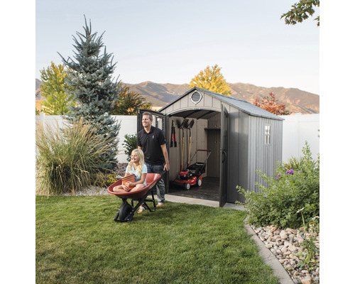 Scéna s kůlnou na nářadí, ve které je uložená sekačka na trávu a zahradní nářadí, zatímco muž tlačí dívku v kolečku.