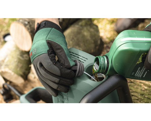 Osoba nalévá olej do řetězové pily. V pozadí jsou vidět kusy dřeva.