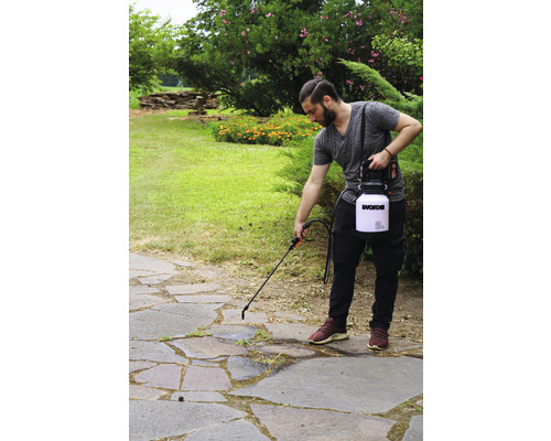 Osoba používá zahradní postřikovač na kamenné terase k hubení plevele. V pozadí jsou keře a zahrada.