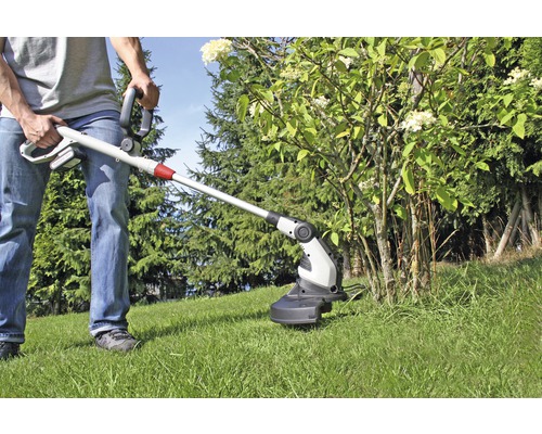 Osoba používá akumulátorový vyžínač na zahradě k sekání trávy pod keřem.