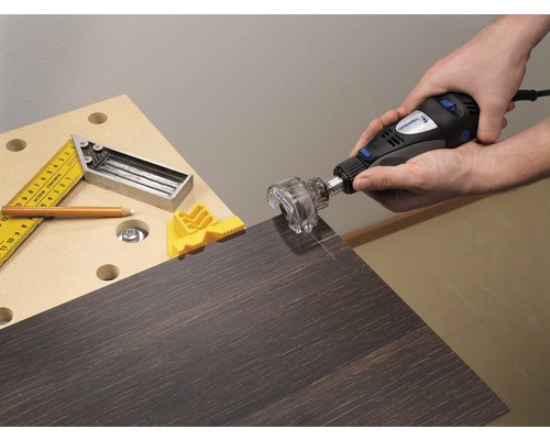 Osoba používá multifunkční nástroj Dremel k řezání kusu dřeva, s úhelníkem, tužkou a žlutým držákem na stole.