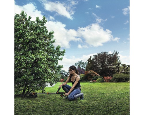 Žena pracuje s akumulátorovou sekačkou na trávu v zahradě na stromě