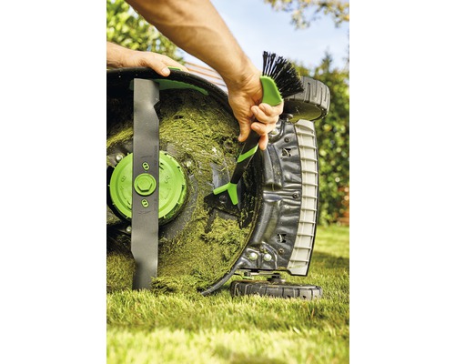 Osoba čistí kartáčem sekačku na trávu od zbytků trávy.