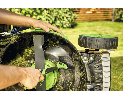 Osoba čistí se kartáčem sekačku na trávu.