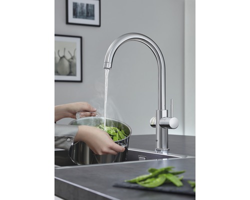 Kuchyňská scéna s dřezovou baterií, u které osoba plní hrnec zeleninou