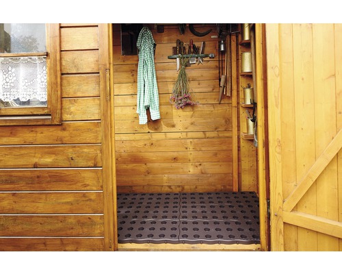 Vnitřní pohled na dřevěný zahradní domek s gumovými rohožemi na podlaze a různým zahradním nářadím na stěnách