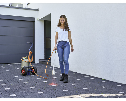 Žena používá plynový hořák k hubení plevele na dlážděné příjezdové cestě před domem s garáží