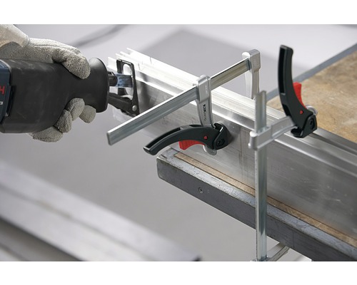 Šavlová pila řeže kovový profil upevněný šroubovými svorkami.