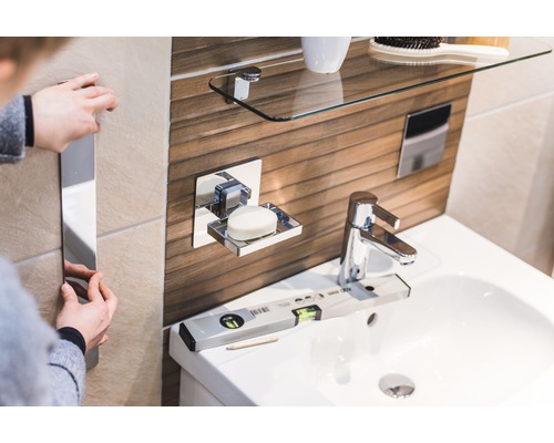 Světlá koupelna s umyvadlem, mýdlenkou, skleněnou policí a osobou, která instaluje zrcadlo