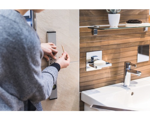 Osoba používá vodováhu a tužku v koupelně k označení přímky na zdi. V pozadí je vidět umyvadlo, vodovodní kohoutek a dávkovač mýdla.