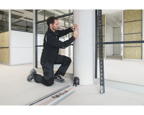 Řemeslník s křížovým liniovým laserem při montáži kovové spodní konstrukce v interiéru.