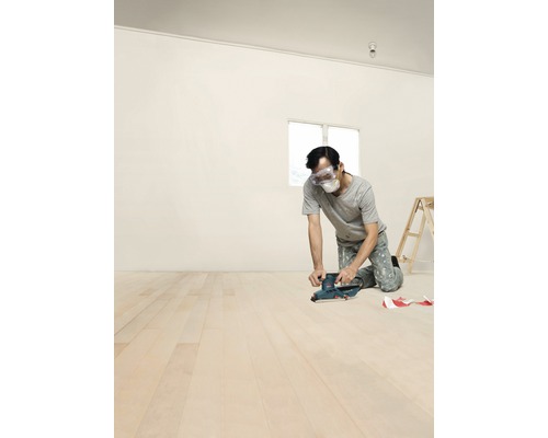Muž brousí dřevěnou podlahu bruskou ve světlé místnosti