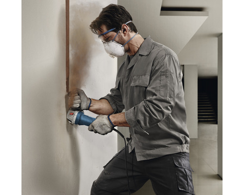 Řemeslník pracuje s drážkovací frézou na zdi. Má ochranné brýle, ústenku a rukavice.