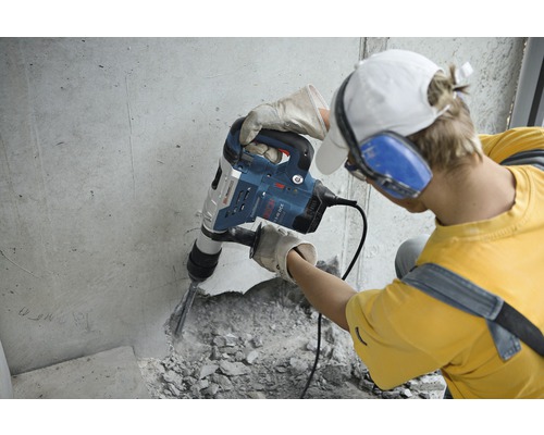 Muž pracuje s bouracím kladivem na betonové zdi. Má na sobě ochranné rukavice a chrániče sluchu.