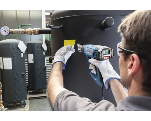 Osoba odstraňuje horkovzdušnou pistolí etiketu z topné olejové nádrže.