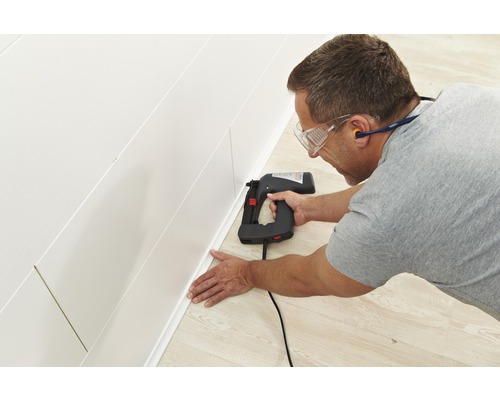 Muž připevňuje soklovou lištu k nástěnnému panelu pomocí elektrické sponkovačky.