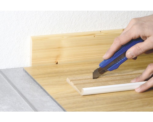 Řezačka koberců řeže bílou podlahovou lištu na dřevěném povrchu.