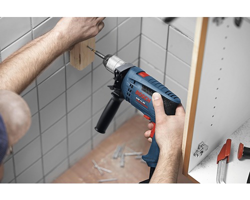 Řemeslník vrtá příklepovou vrtačkou do kusu dřeva na zdi. V pozadí je vidět skříňka. Na podlaze leží šrouby.