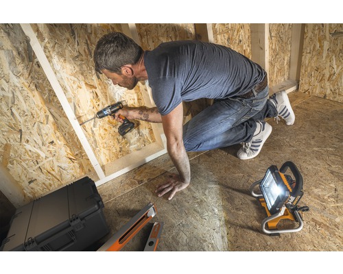 Řemeslník pracuje s akumulátorovým šroubovákem v hrubé stavbě ze dřeva, osvětlené stavebním reflektorem.