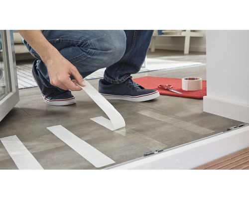 Muž pokládá podlahovou krytinu pomocí lepicí pásky.