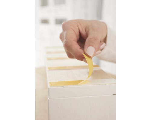 Ruka odstraňuje ochrannou fólii z lepicí pásky na bílém předmětu.