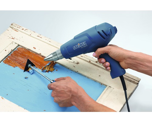 Osoba odstraňuje zbytky barvy z dveří horkovzdušnou pistolí a špachtlí