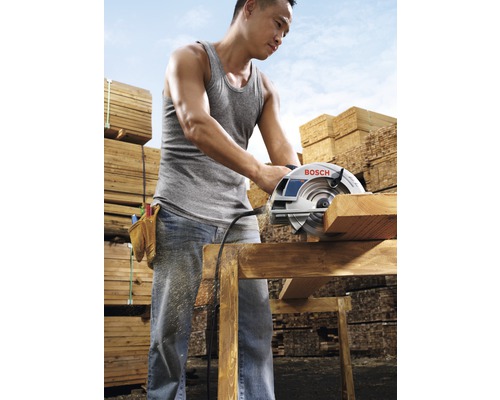 Muž řeže dřevěnou desku kotoučovou pilou, v pozadí hromady dřeva.