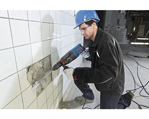 Řemeslník s ochrannými brýlemi, chrániči sluchu a ochrannou helmou pracuje s vrtacím kladivem na obkládané stěně