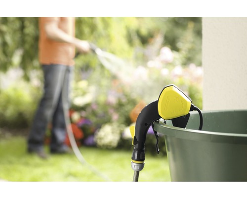 Zahradní scéna s zavlažovací tyčí připevněnou k vědru a osobou v pozadí zalévající zahradu.