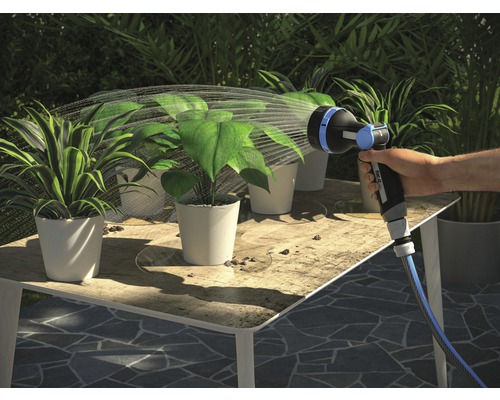 Ruka zavlažuje rostliny zahradní hadicí na stole.