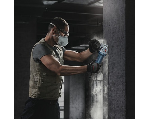 Muž s ochrannou maskou a brýlemi pracuje s úhlovou bruskou na betonové zdi.
