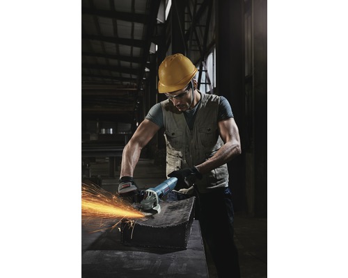 Pracovník s ochrannou helmou a brýlemi používá úhlovou brusku na kusu kovu, což způsobuje jiskry.