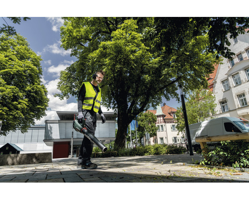 Muž s bezpečnostní vestou a sluchátky používá venku foukač listí.