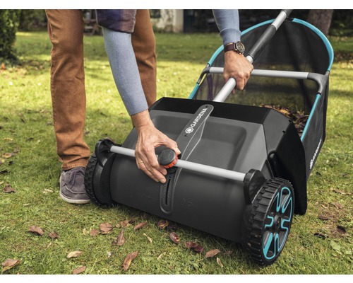 Osoba obsluhuje sběrač trávy Gardena na trávníku s listím.