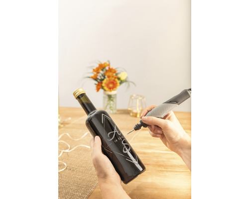 Osoba gravíruje skleněnou láhev iniciálami a datem pomocí gravírovacího nástroje Dremel.