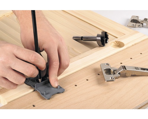 Osoba označuje pozici pantu pomocí šablony a tužky na dřevěné desce, vedle Forstnerova vrtáku a pantu.