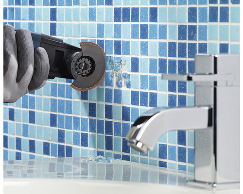 Pracovník v rukavicích odstraňuje dlaždice multifunkčním nástrojem v koupelně