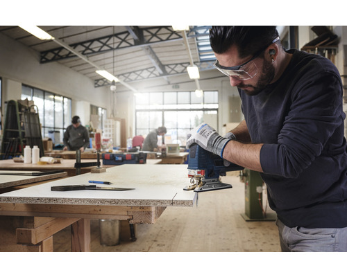 Muž řeže obrobek přímočarou pilou v dílně.