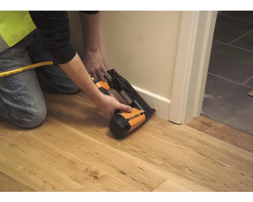 Osoba připevňuje soklovou lištu ke zdi pomocí pneumatické sponkovačky.