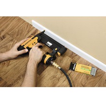 Osoba používá pneumatickou sponkovačku k upevnění podlahové lišty. Vedle leží balení odpovídajících hřebíků.