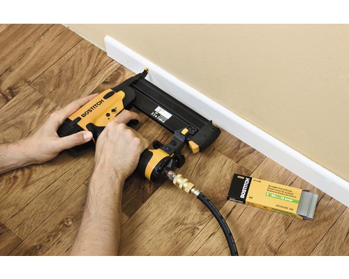 Osoba používá pneumatickou sponkovačku k upevnění podlahové lišty. Vedle leží balení odpovídajících hřebíků.