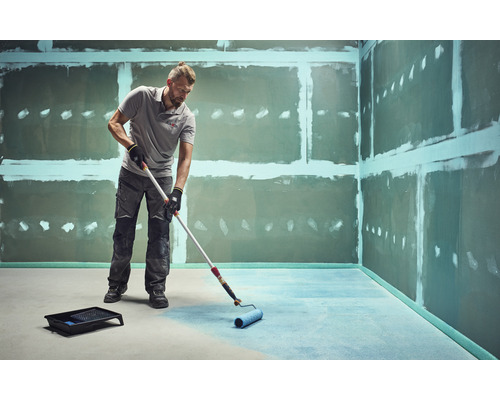 Muž natírá podlahu válečkem v nedokončené místnosti.